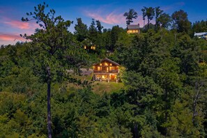 A two-story wooden cabin surrounded by dense forest, with a clear view of the property.