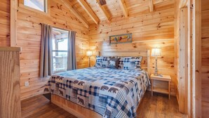 A wooden cabin bedroom with a bed, two bedside tables, a window with curtains, and a wooden ceiling.