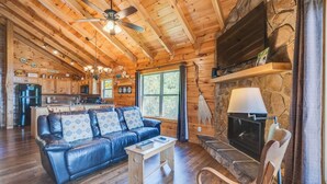 A cozy cabin interior with a leather sofa, wooden coffee table, stone fireplace, and a kitchen area visible through an open door.