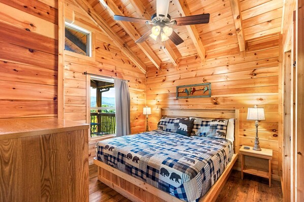 A wooden cabin bedroom with a bed, a ceiling fan, a nightstand, and a window with a view.
