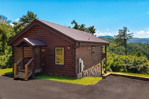 Eine kleine Holzhütte mit Veranda und Treppe, umgeben von üppigem Grün und Bergen im Hintergrund.