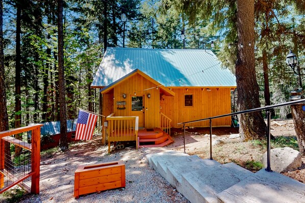 Una cabana de fusta amb teulada metàl·lica, envoltada d'arbres, i amb un porxo que té uns escalons que porten a una porta.