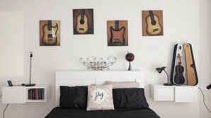 Quarto com cama, mesa de cabeceira com livros, guitarra presa à parede e um objeto decorativo de metal sobre a cama.