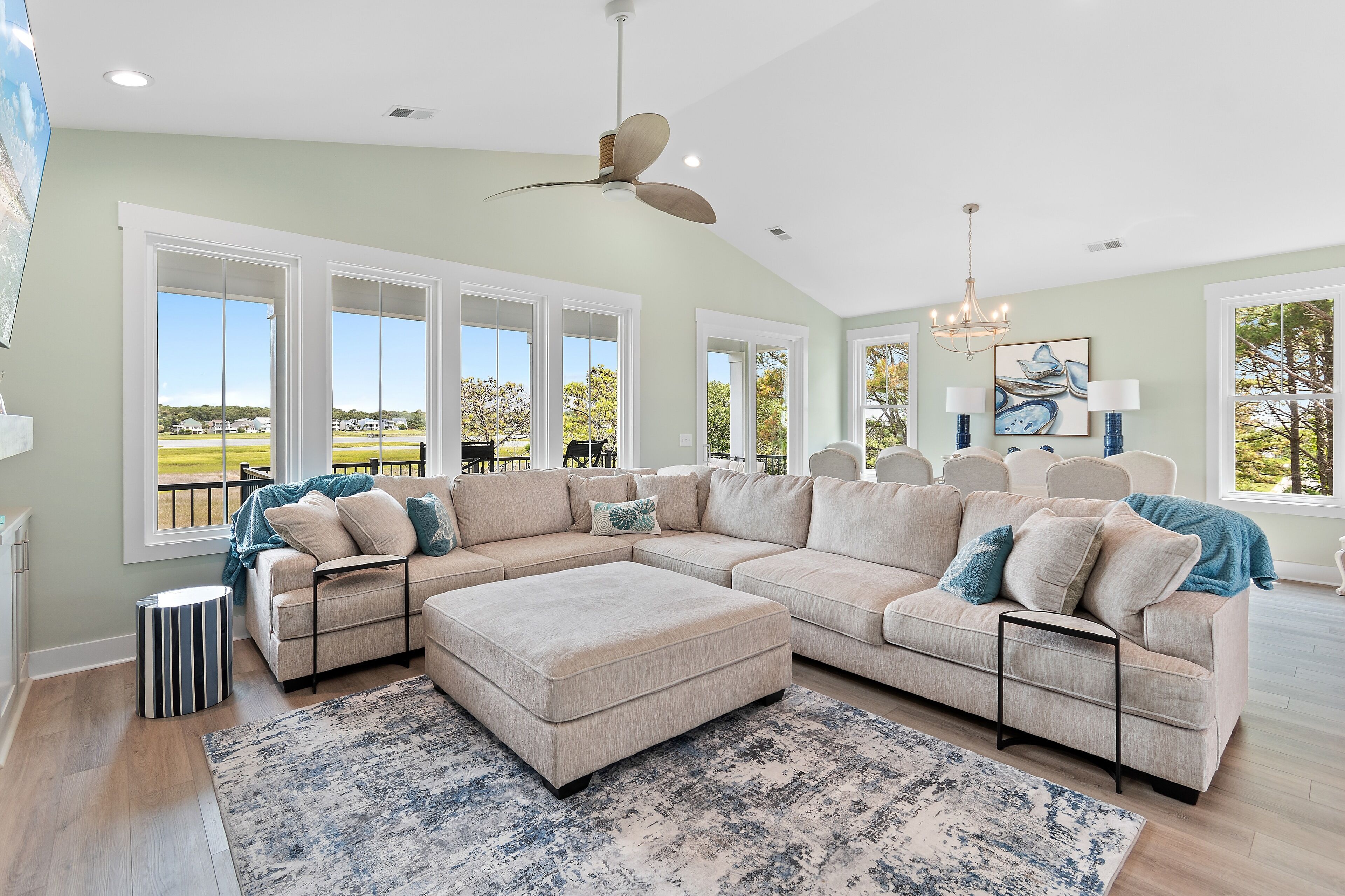 A spacious living room with a large sectional sofa, a matching ottoman, and a patterned rug. There are multiple windows providing natural light and views of the outdoors.