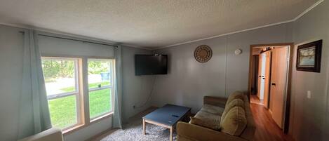 A living room with a sofa, a coffee table, and a TV mounted on the wall. There is a window with curtains, a door, and a rug on the floor.