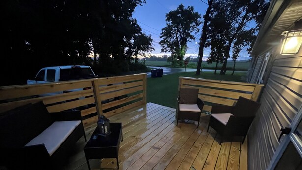 Una terraza de madera con mobiliario exterior y una pequeña mesa, rodeada de árboles y un césped.