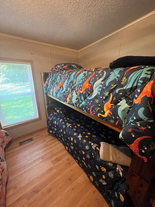 Una habitación pequeña con una cama litera decorada con un edredón de temática de dinosaurios, una ventana con persianas y suelo de madera.