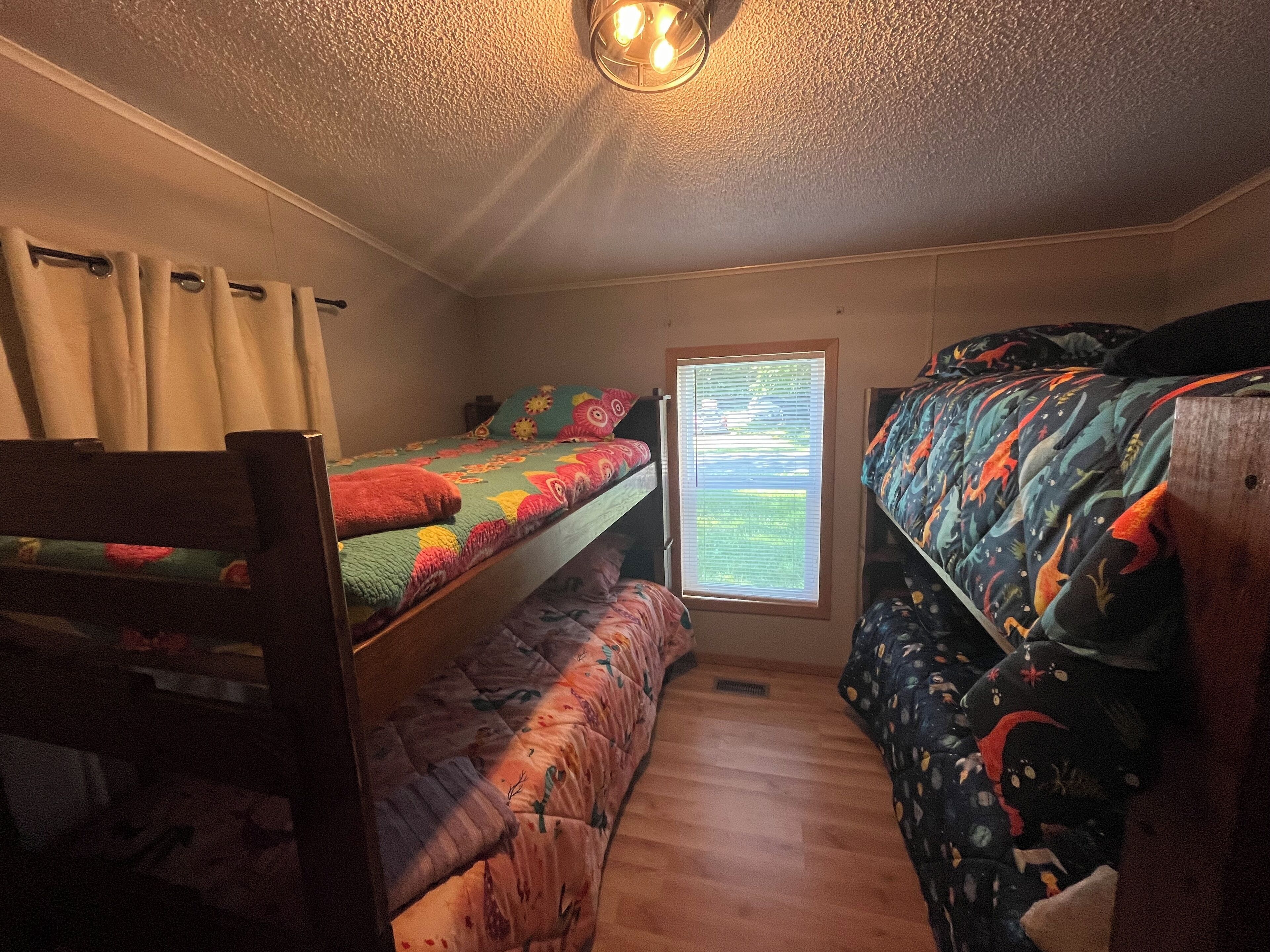 A small, cozy room with two bunk beds, a window with blinds, and a ceiling light fixture.