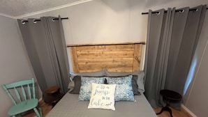 Un dormitorio con cabecera de madera, cortinas grises, una silla color turquesa y una cama con almohadas estampadas.