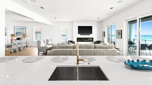 A modern living room with a large sofa, a dining area with a table and chairs, a kitchen with a sink and countertop, and a balcony with a view of the ocean. - Luxurious Beachfront Retreat: Pool, Spa, Chefs Kitchen (Destin)