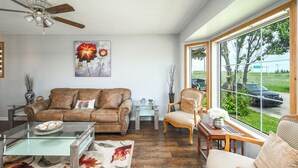 A living room with a brown leather sofa, a glass coffee table, and a floral rug. There is a window with a view of a road and greenery outside.
