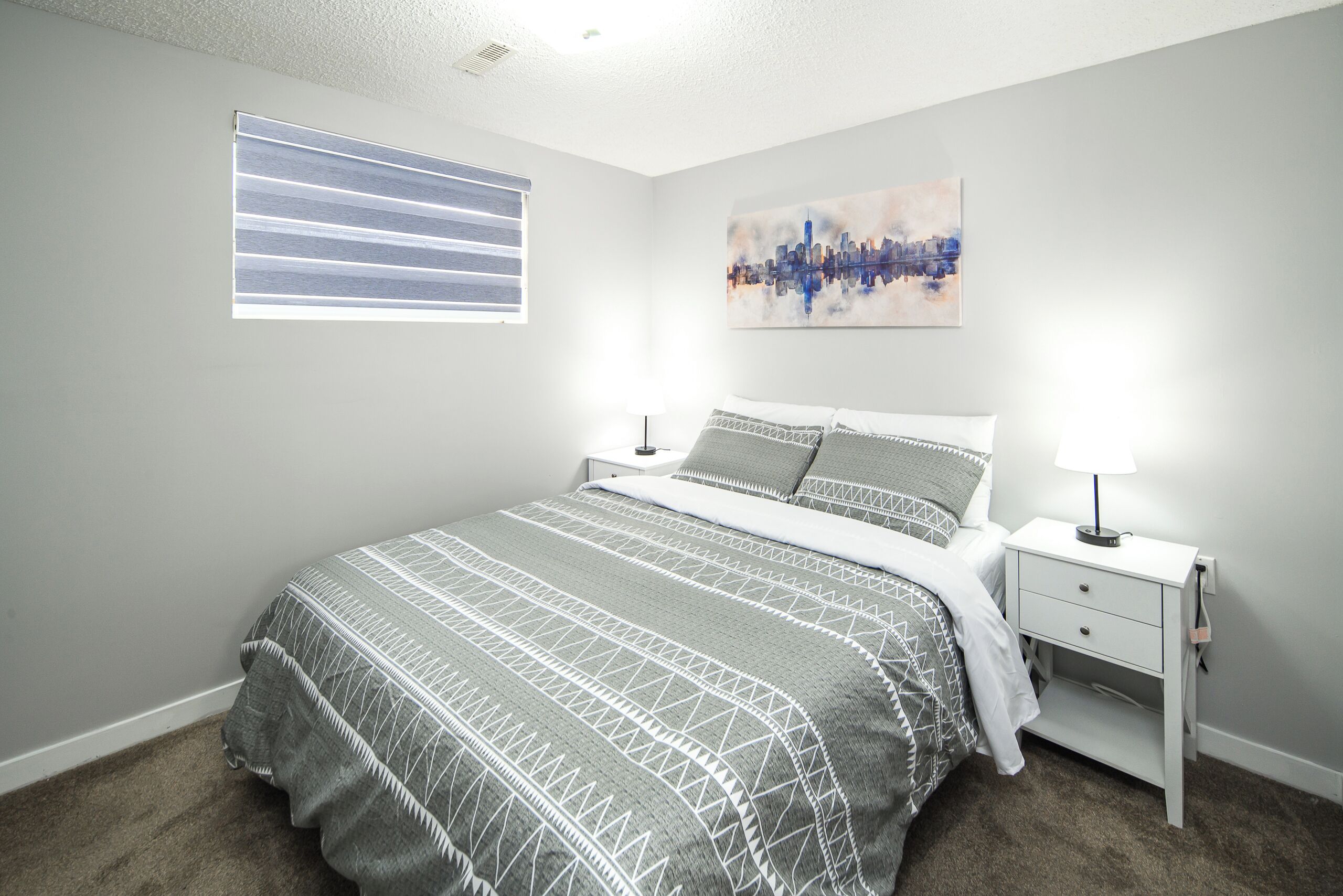 Une chambre à coucher avec un lit, une table de chevet, une lampe, une fenêtre avec des stores et un tableau représentant un paysage urbain accroch é au mur.
