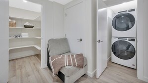 A laundry room with a washing machine and dryer, a gray couch with a blanket, and a shelving unit with baskets and towels.