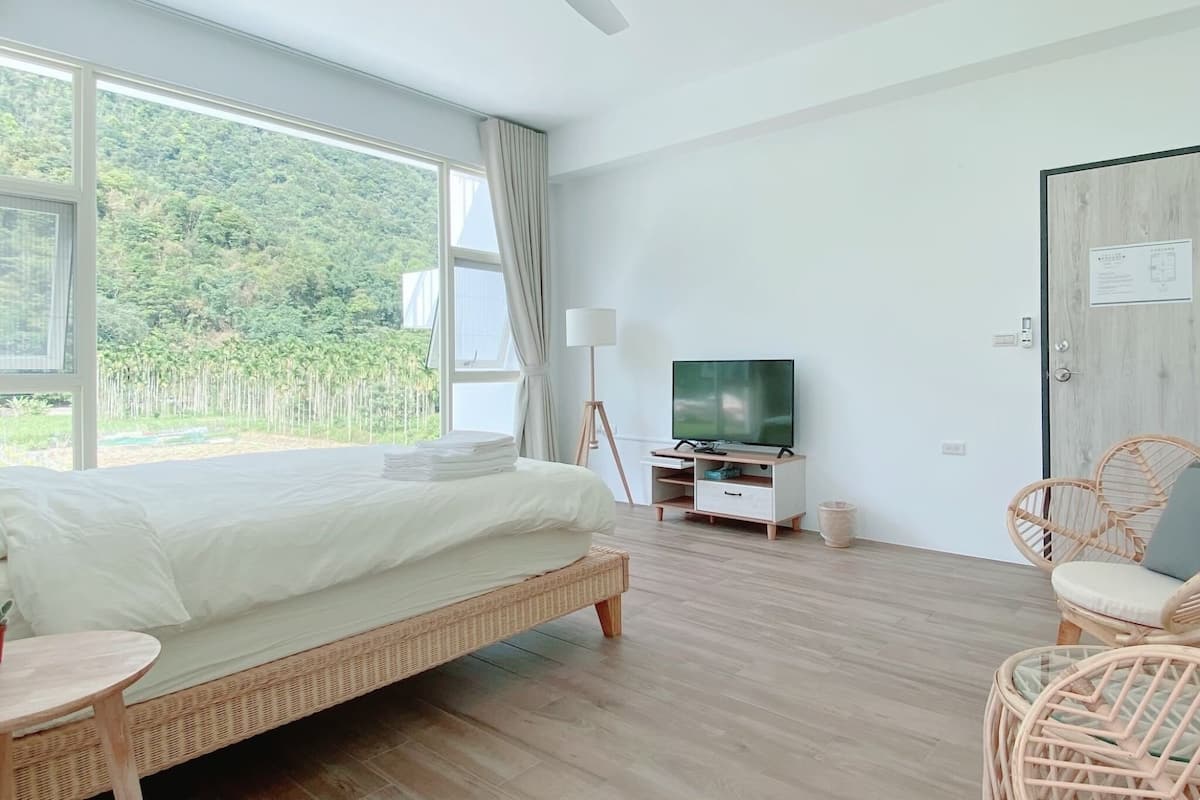 A bedroom with a bed, a TV, a lamp, a chair, and a view of greenery outside.