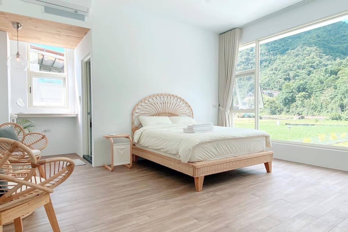 A bedroom with a large window, a bed, a bedside table, and a chair.