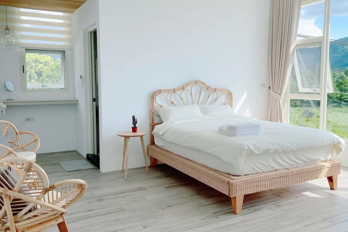A room with a bed, wicker chairs, a small table, and a window with a view of greenery.
