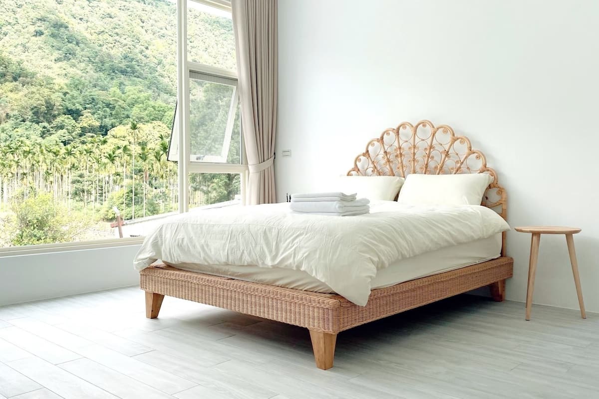 A bedroom with a wicker bed, white bedding, and a view of a forest through a large window.