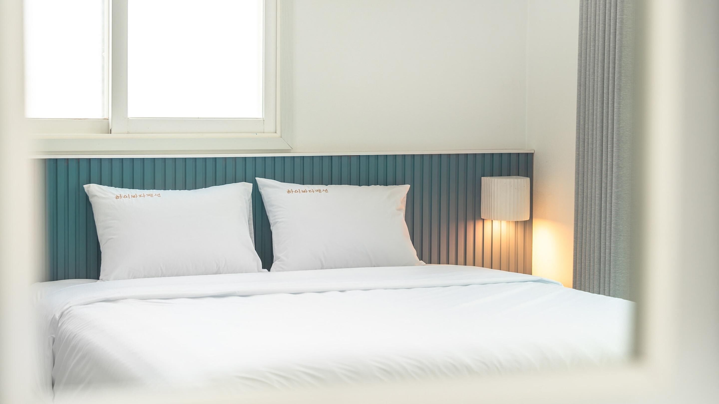 Ein Hotelzimmer mit einem Bett, zwei Kissen, einem Nachttisch mit Lampe und einem Fenster mit Jalousien.