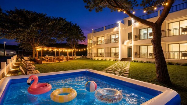 An outdoor swimming pool with inflatable toys, a well-lit building with balconies, and a paved patio area. - Taean Yeonpo Haibada Pension (Taean)