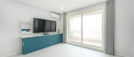 A modern, minimalist room with a large window, a flat-screen TV mounted on a blue cabinet, and a small bedside table.