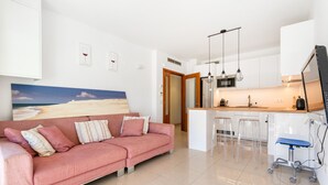 Un salon moderne avec un canapé rose, une cuisine aux meubles blancs et un grand tableau représentant une scène de plage.
