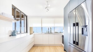 A modern kitchen with white cabinets, a stainless steel refrigerator, and a window with a view of trees and sky. - BANNOW7 (Alexandra Headland)