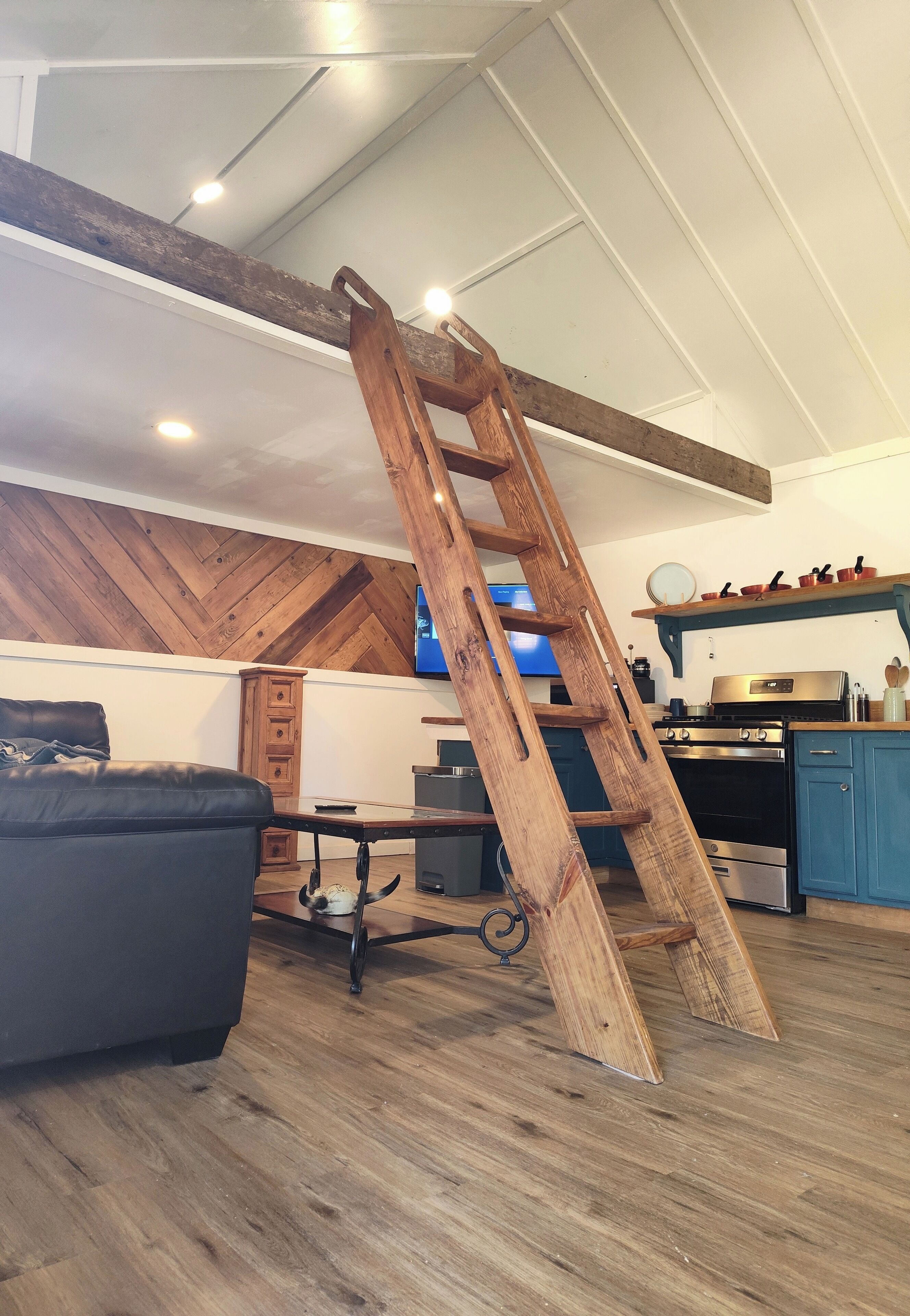 A loft-style living area with exposed wooden beams, a wooden ladder, a black leather couch, a kitchen with blue cabinets, and a television on the wall.