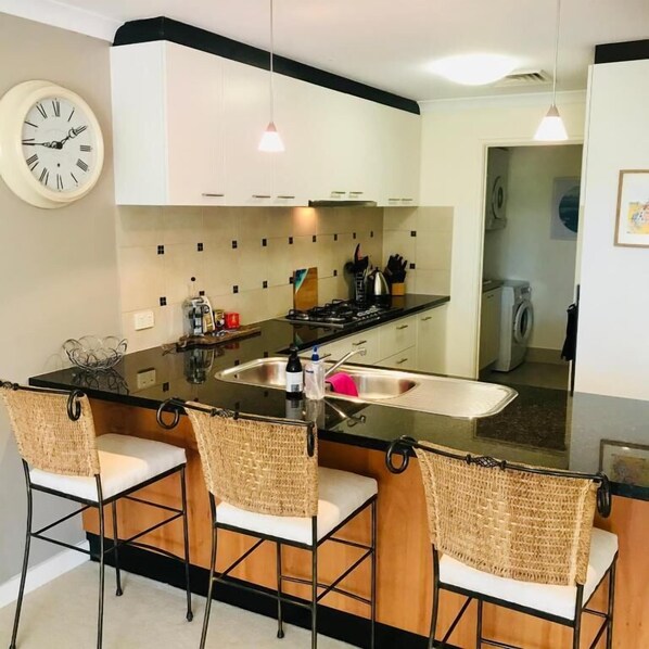 A modern kitchen with a central island, bar stools, a clock, and a washing machine in the background.
