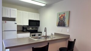 A compact kitchen with white cabinets, a black microwave, a stove, and a sink. There are two bar stools and a painting on the wall.