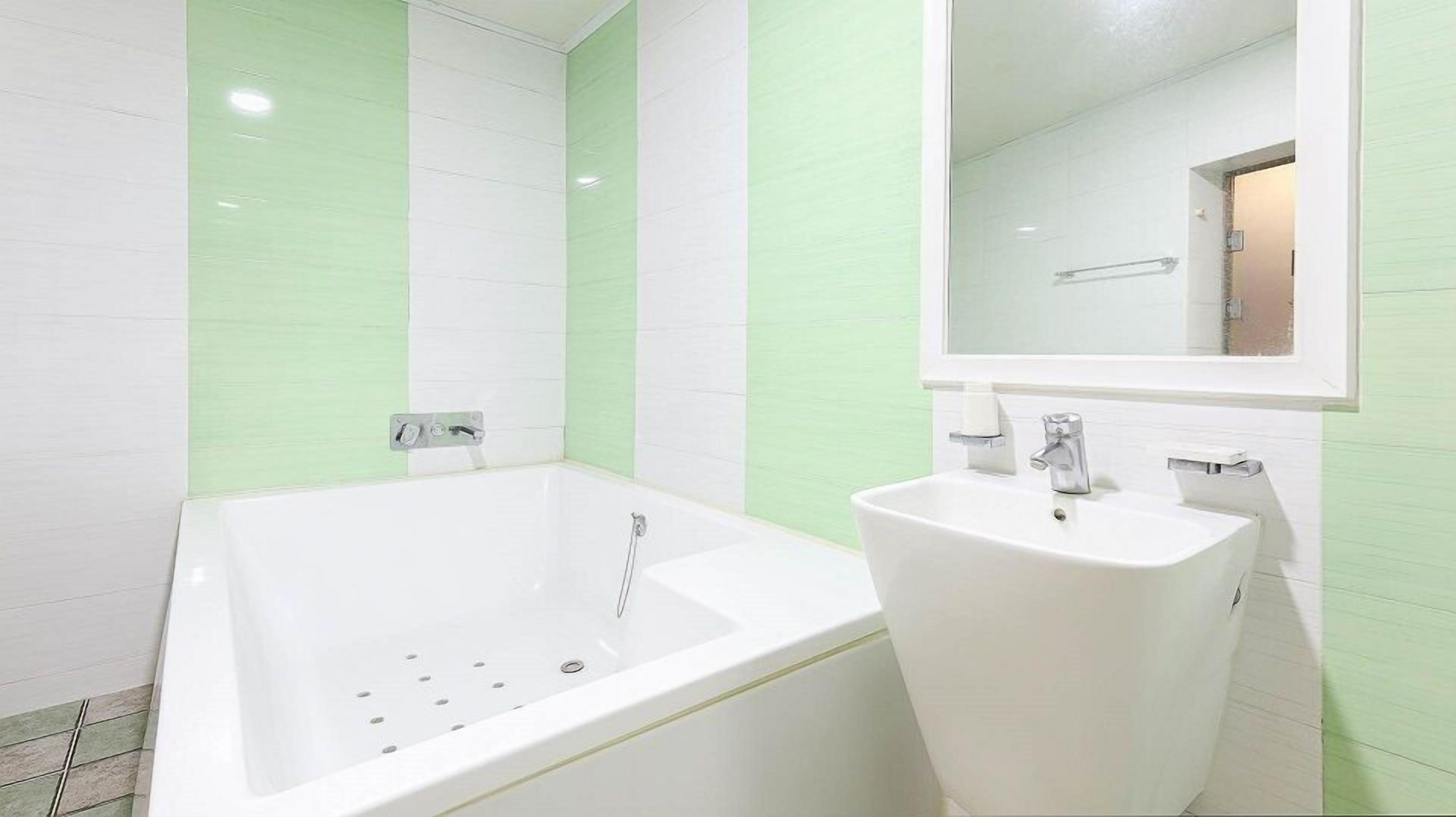 Un baño con bañera y lavamanos blancos, frente a una pared con azulejos verdes y blancos.