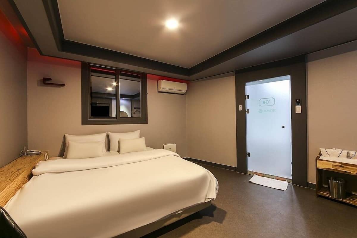 A hotel room with a bed, a desk, a mirror, an air conditioning unit, and a door with a number.