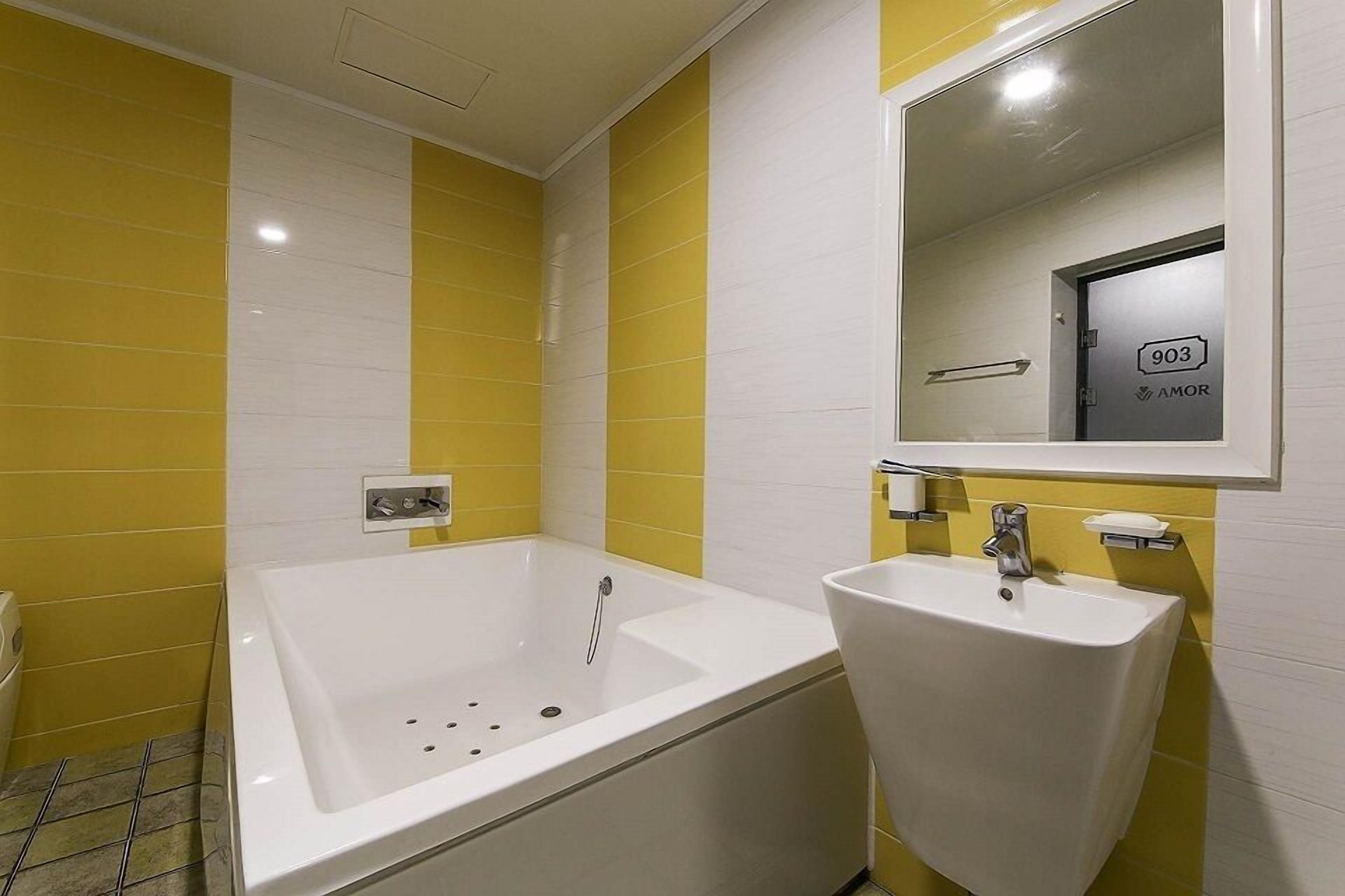 A bathroom with yellow and white tiles, a bathtub, a sink, and a mirror reflecting a door with the number 903 and the word "AMOR".