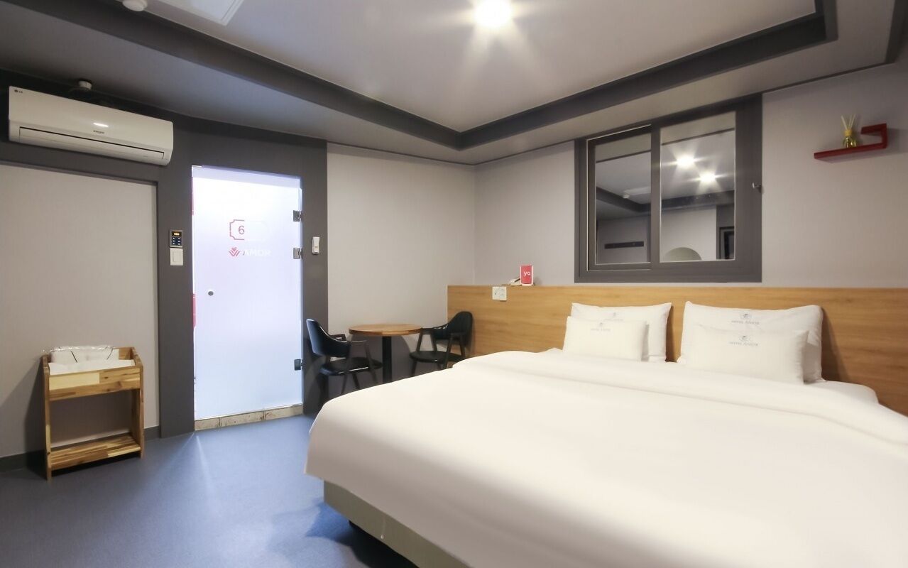 A hotel room with a large bed, a desk, a chair, a small table, an air conditioning unit, and a door with a number sign.