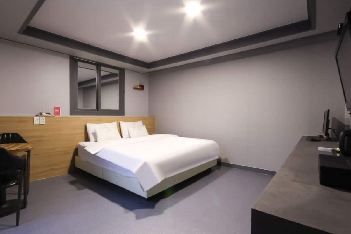 A modern hotel room with a bed, a desk, a chair, a small table, a mirror, and a clock on the wall.