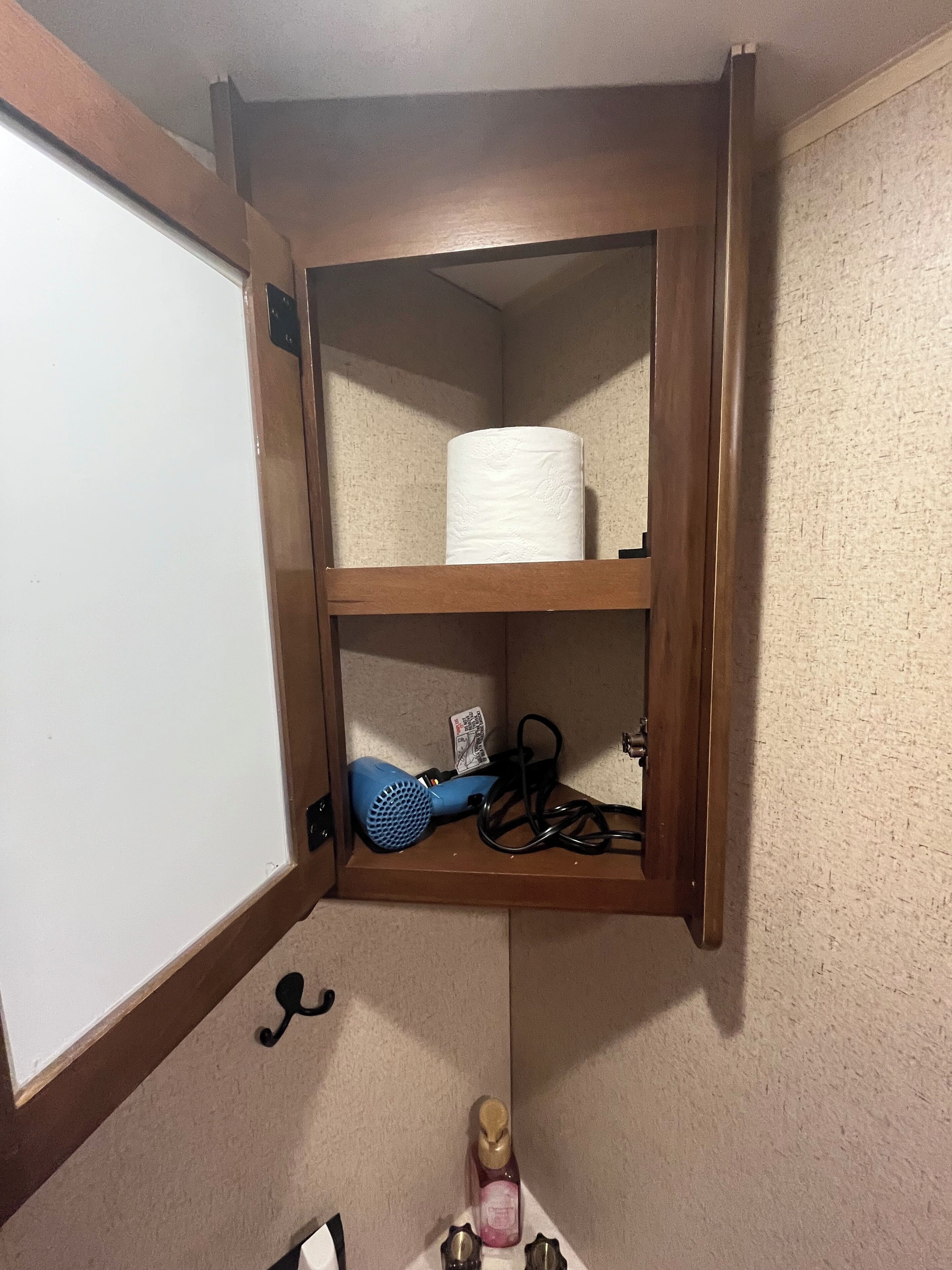A wooden cabinet with two shelves, one holding a roll of toilet paper and the other containing a blue hairdryer and a bottle.