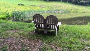 Un banc doté d’un dossier et d’accoudoirs, placé sur une pelouse près d’un étang bordé d’arbres.