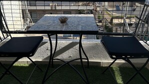 En balkong med ett bord och två stolar, med utsikt över en stadssiluett med byggnader och en klar himmel.