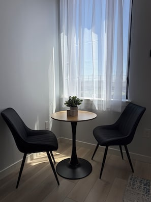 A room with a round table, two chairs, a potted plant, sheer curtains, and a view through the window.