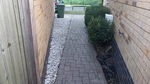 Un estrecho sendero de ladrillo que lleva a una zona vallada, con un bote verde para basura y una cerca.