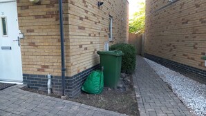 Un edificio de ladrillo con un sendero pavimentado, un bote de basura verde y una puerta blanca.