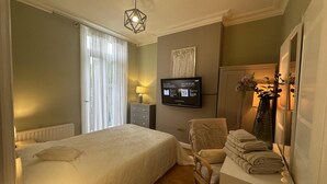 Una habitación de hotel con cama, una silla, un escritorio con lámpara, televisor y ventana con cortinas.