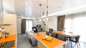 A modern hotel room with a dining area, a sofa, a TV, and a fruit bowl on the island.