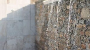 A stone wall with water flowing over it, a tiled floor, and a building in the background.