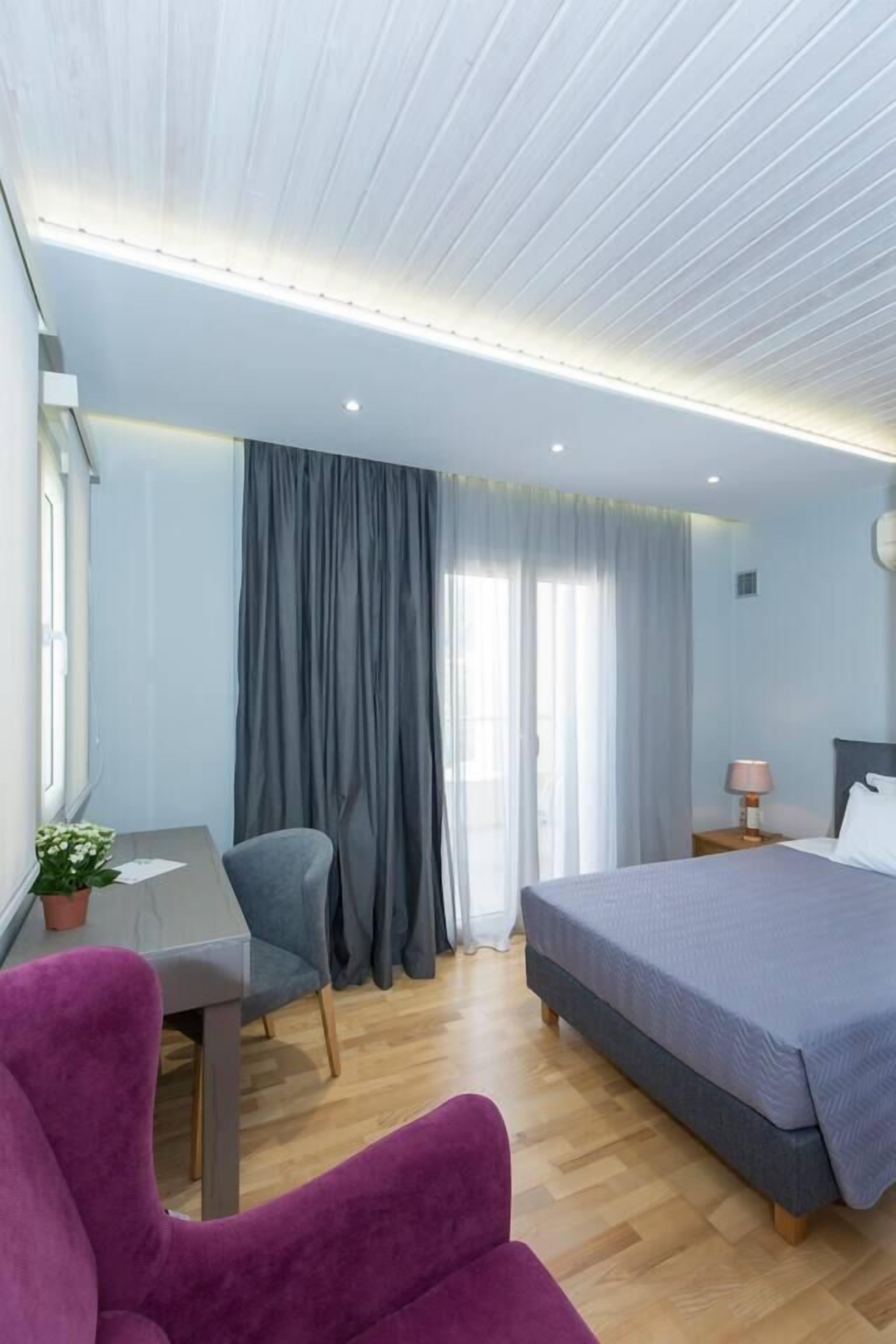 A modern hotel room with a wooden floor, a bed with a grey duvet, a desk with a chair, and a purple armchair.