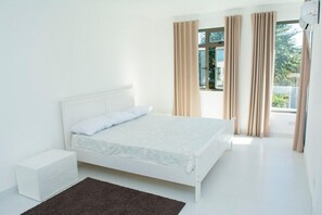 Un dormitorio minimalista con una cama individual, una mesita de noche, una ventana con cortinas y vistas al verdor exterior.