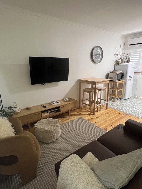 A modern living room with a flat-screen TV mounted on the wall, a wooden dining table with chairs, a sofa, and a small kitchen area with a microwave and refrigerator.