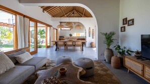 A spacious living area with a sofa, ottomans, a wooden coffee table, and a TV on a wooden cabinet. There is a kitchen in the background with a dining table and chairs. - Villa Rumiano II (Ubud)
