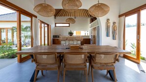 A spacious dining area with a large wooden table and woven chairs, large windows, and a view of the outdoors. - Villa Rumiano II (Ubud)