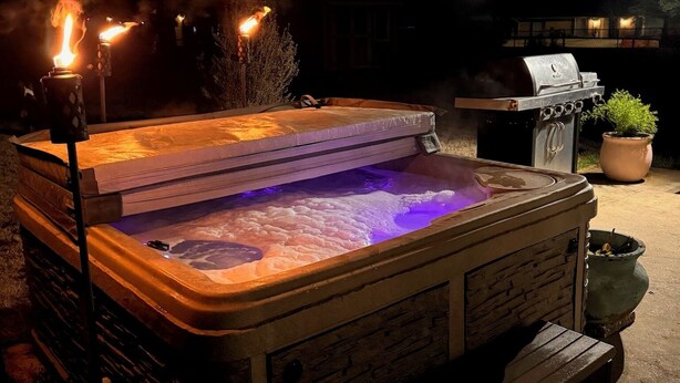 A hot tub with a wooden exterior and a glass top, surrounded by a patio area with a grill and potted plants.