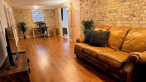 A spacious living room with a leather sofa, wooden flooring, and a brick wall.